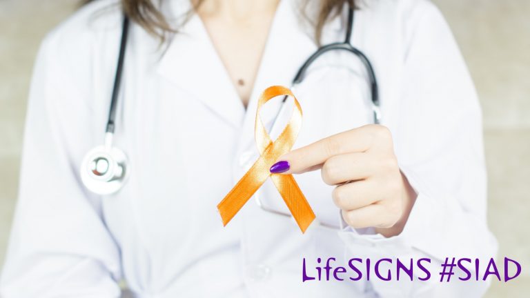 A nurse or doctor in a white coat holds an orange awareness ribbon.
