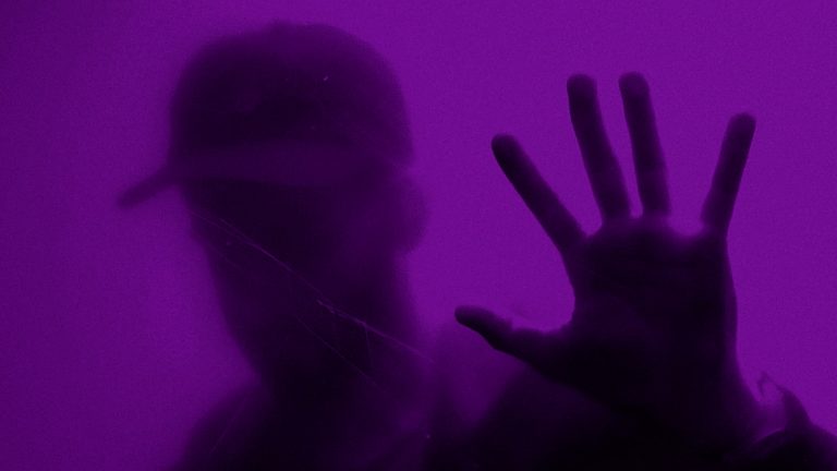 A person in a cap seemingly behind a purple glass wall with their hand up.